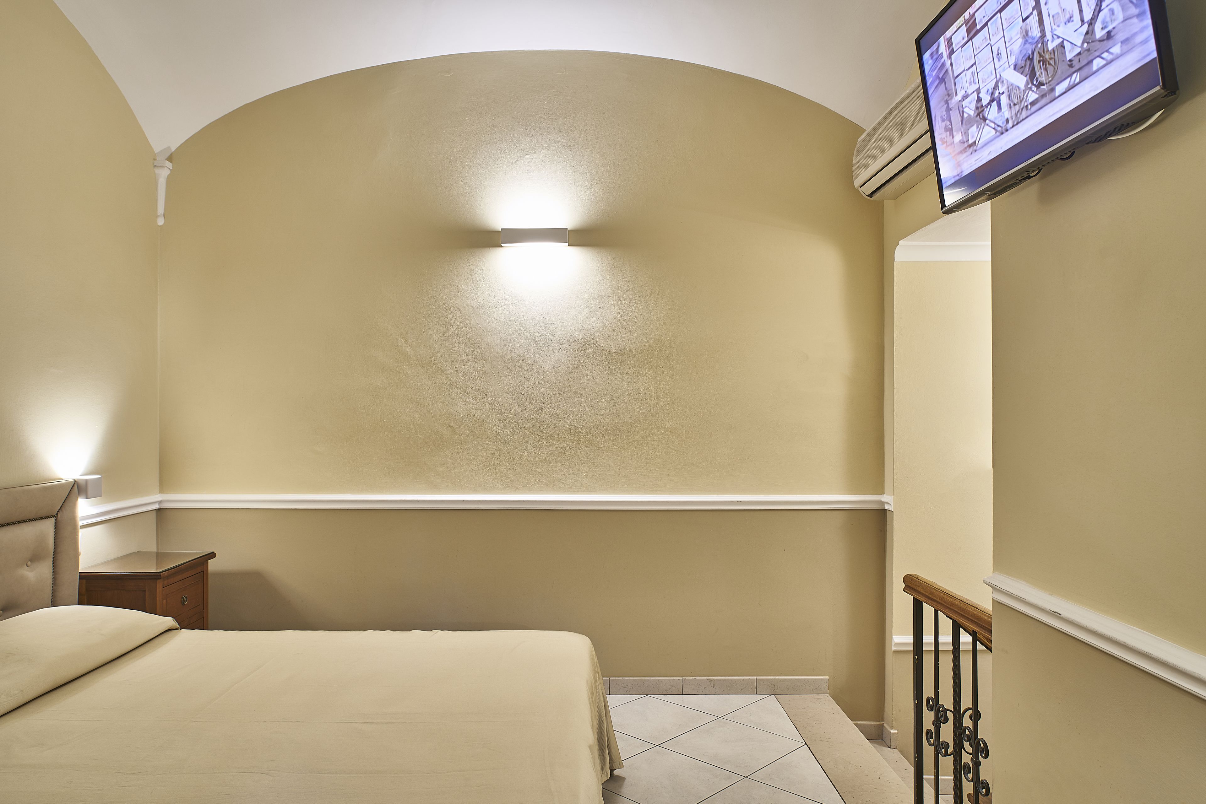 Una habitación de hotel con una cama, una mesita de noche, un televisor montado en la pared y suelo de baldosas.