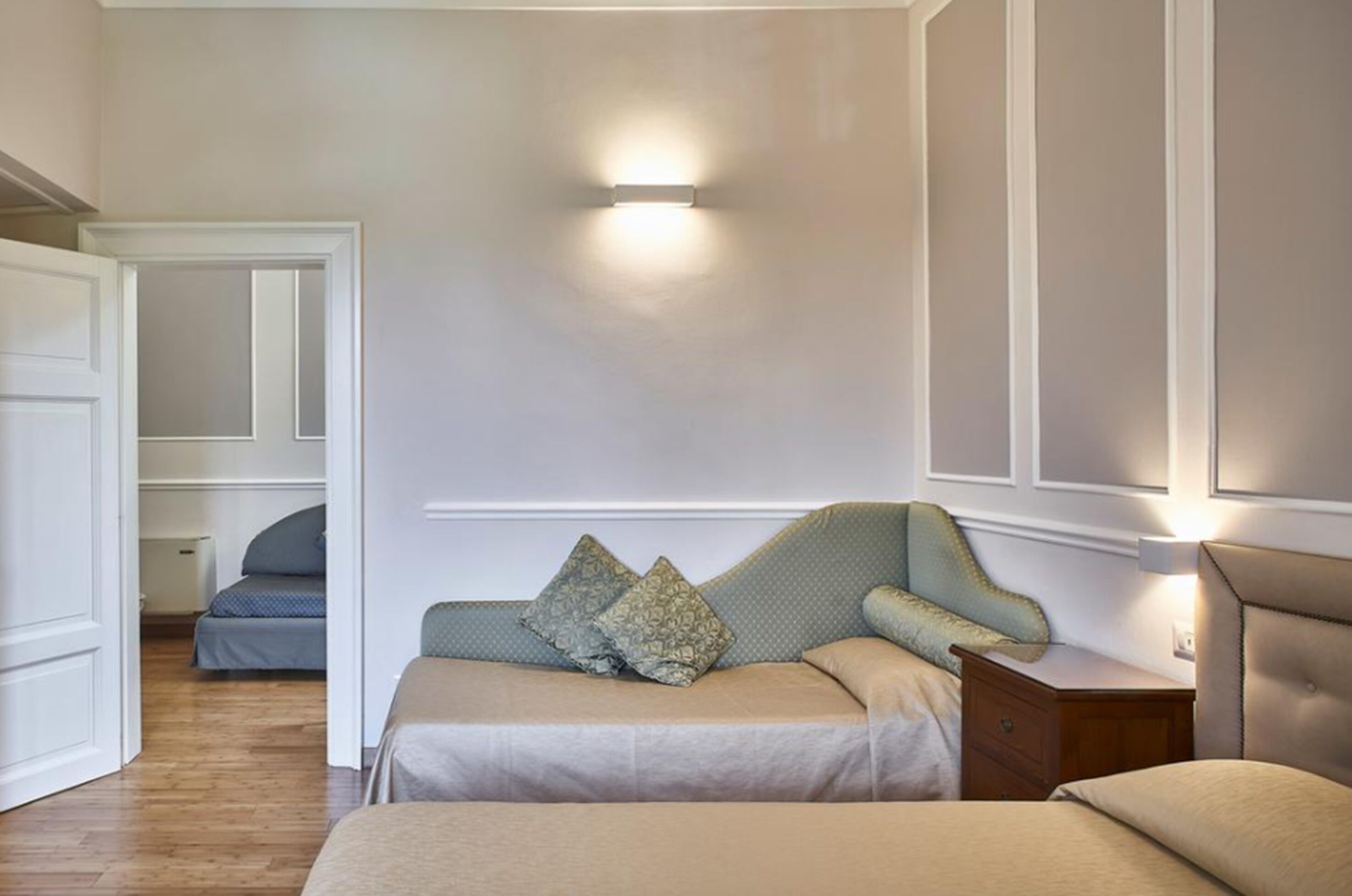 A bedroom with a bed, a nightstand, a wall-mounted light, and a door leading to another room.