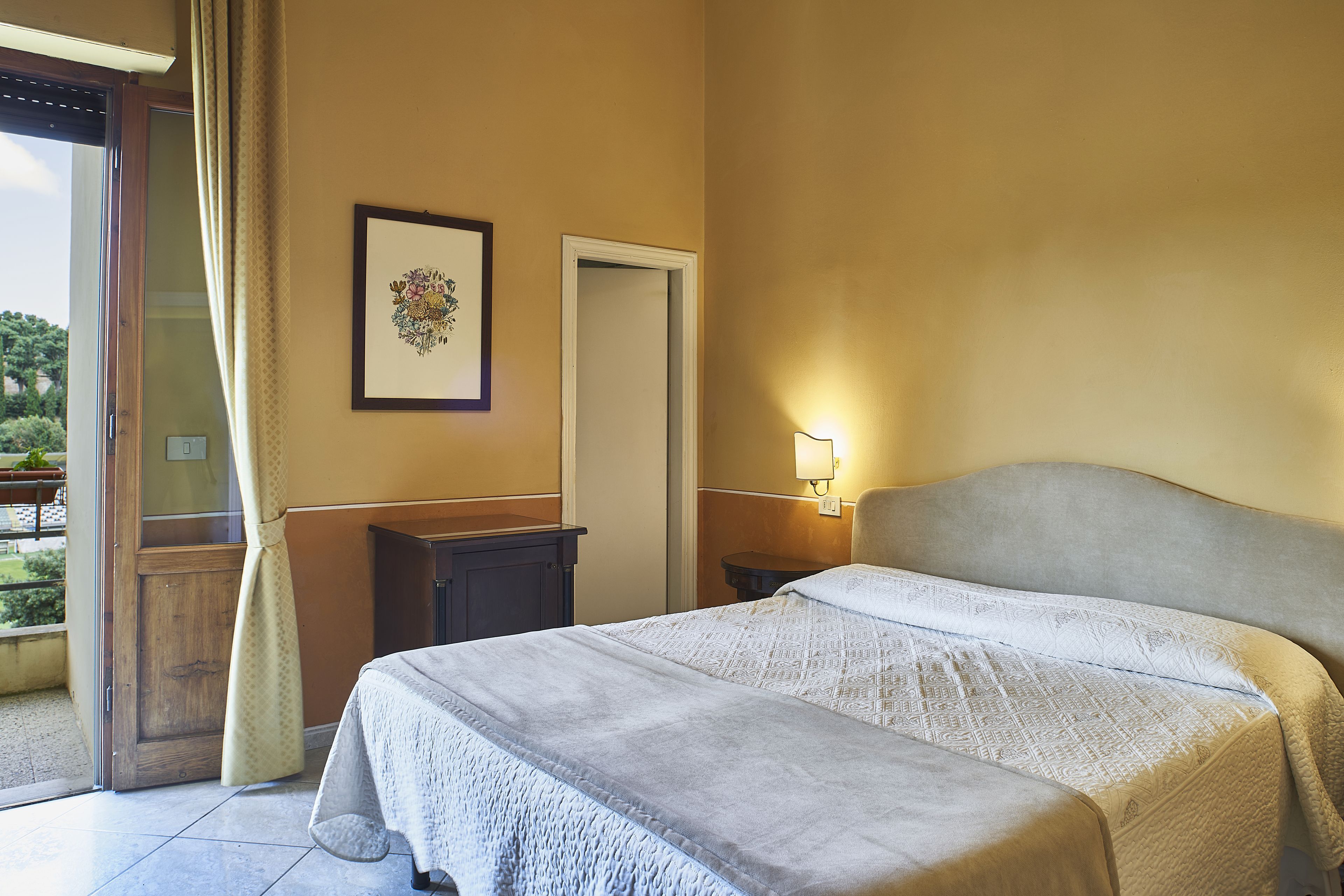 A hotel room with a bed, a bedside table, a lamp, a door, and a window with curtains.