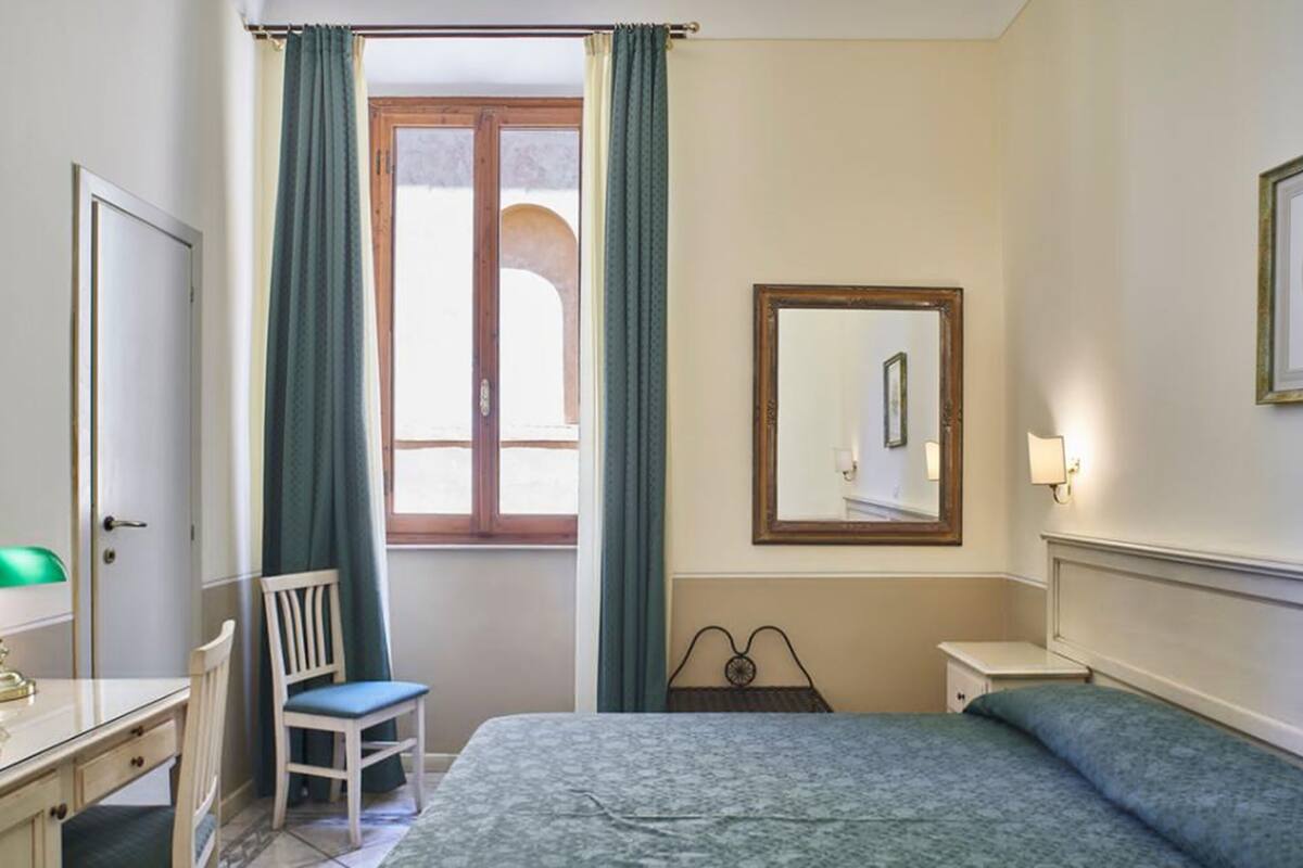 Habitación de hotel con cama, escritorio, silla, ventana con cortinas, espejo y mesita de noche.