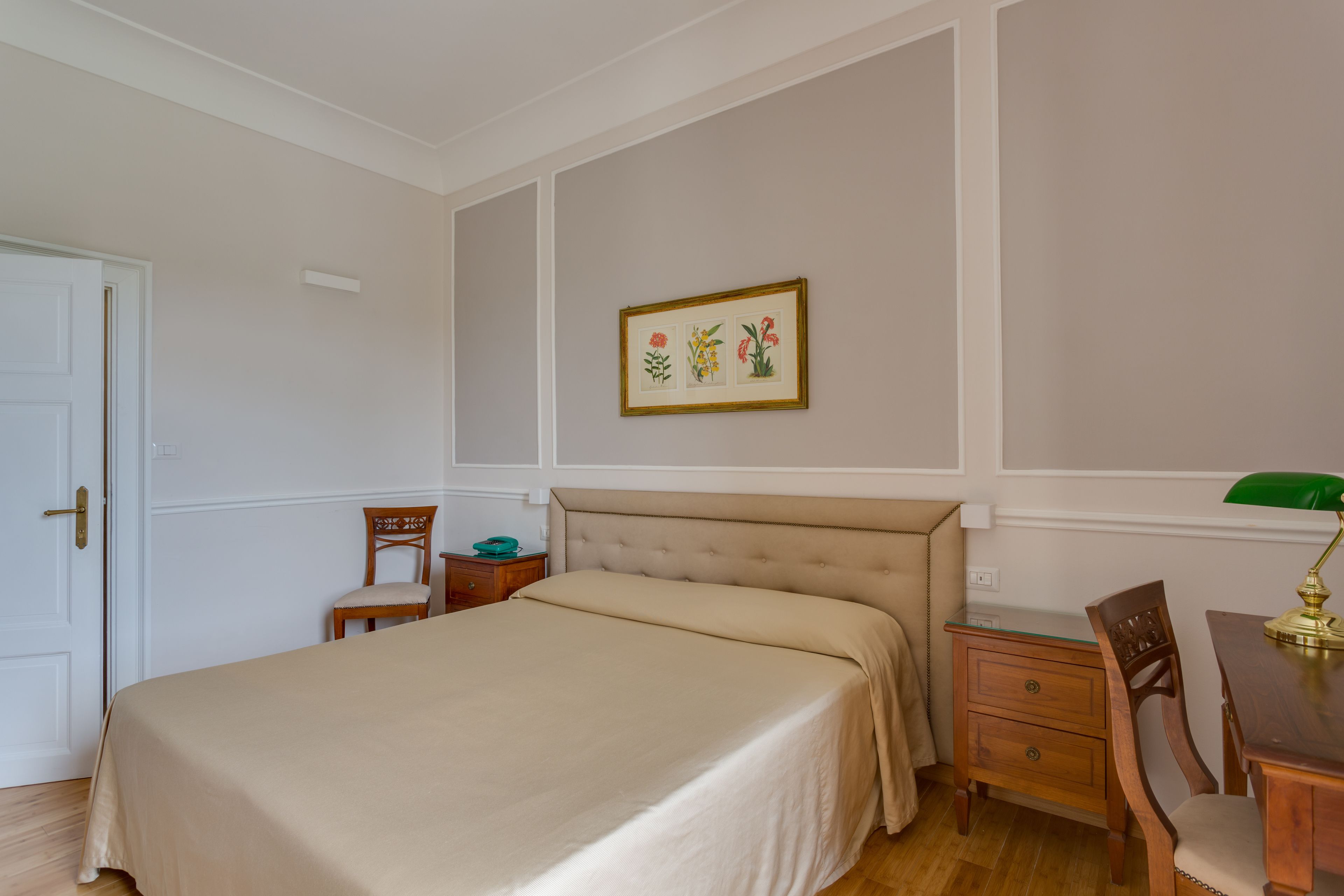 A bedroom with a bed, a nightstand, a chair, a desk, and a framed botanical illustration on the wall.