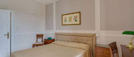 A bedroom with a bed, a nightstand, a chair, a desk, and a framed botanical illustration on the wall.