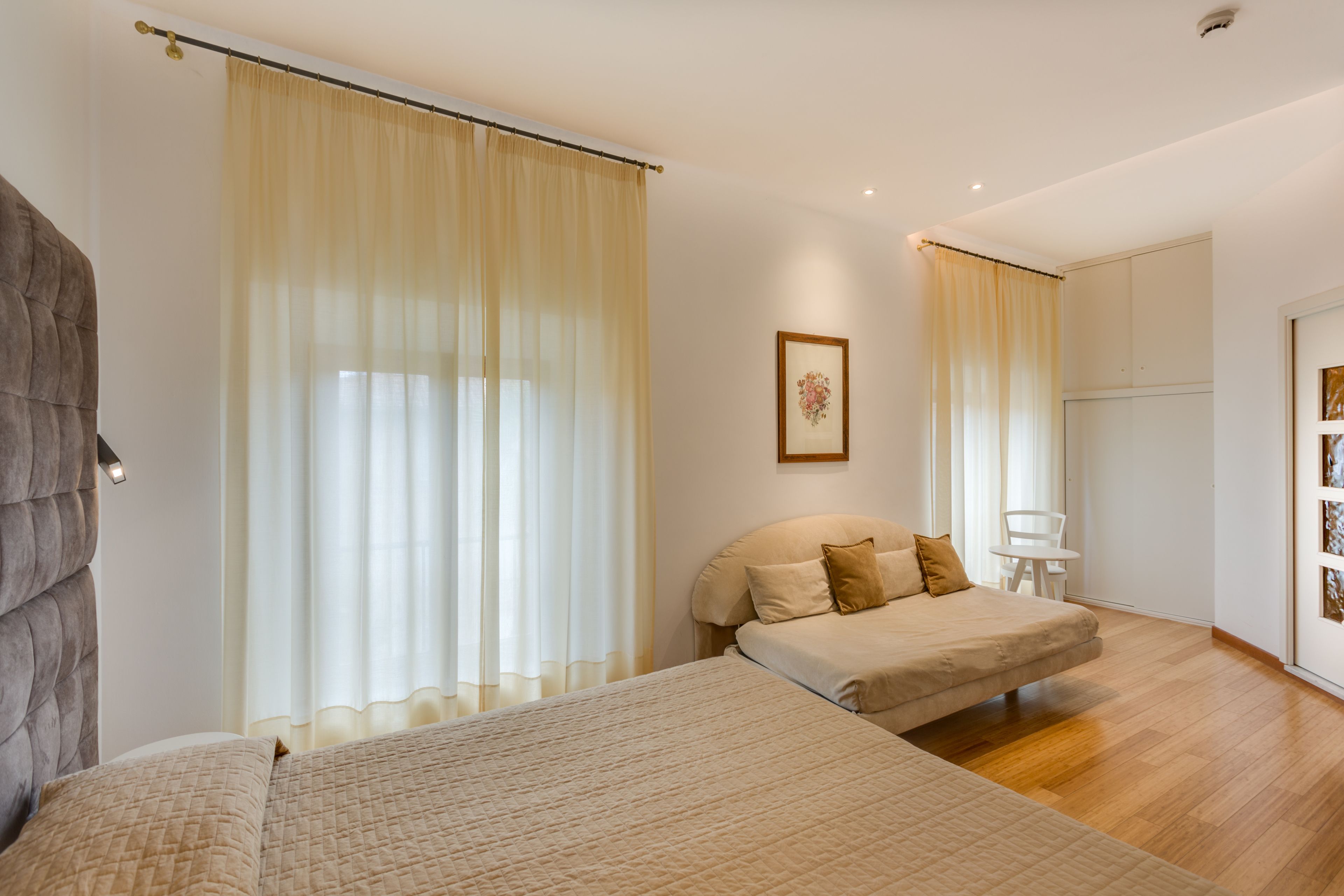 Quarto com duas camas, uma janela grande com cortinas translúcidas, um quadro na parede e uma cadeira branca perto da janela.