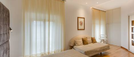 A bedroom with two beds, a large window with sheer curtains, a framed picture on the wall, and a white chair by the window.