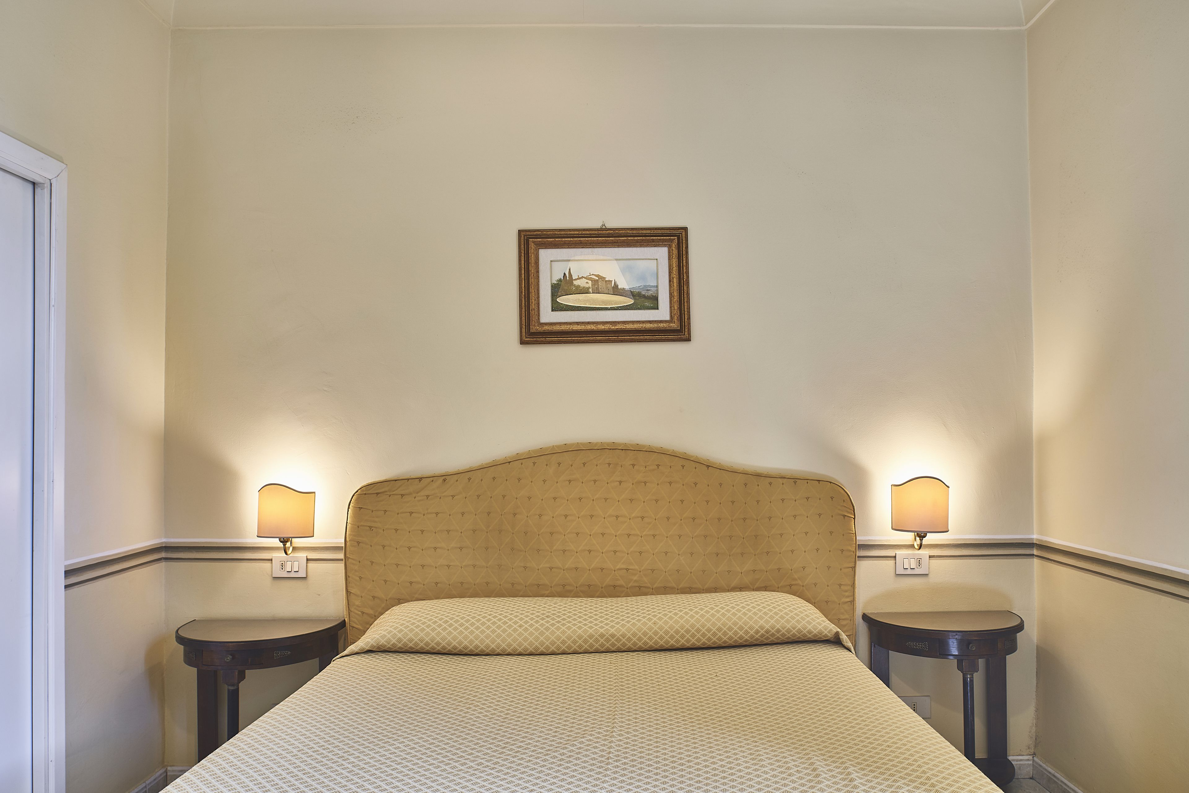A hotel room with a bed, two bedside tables, a wall-mounted lamp, and a framed picture on the wall.