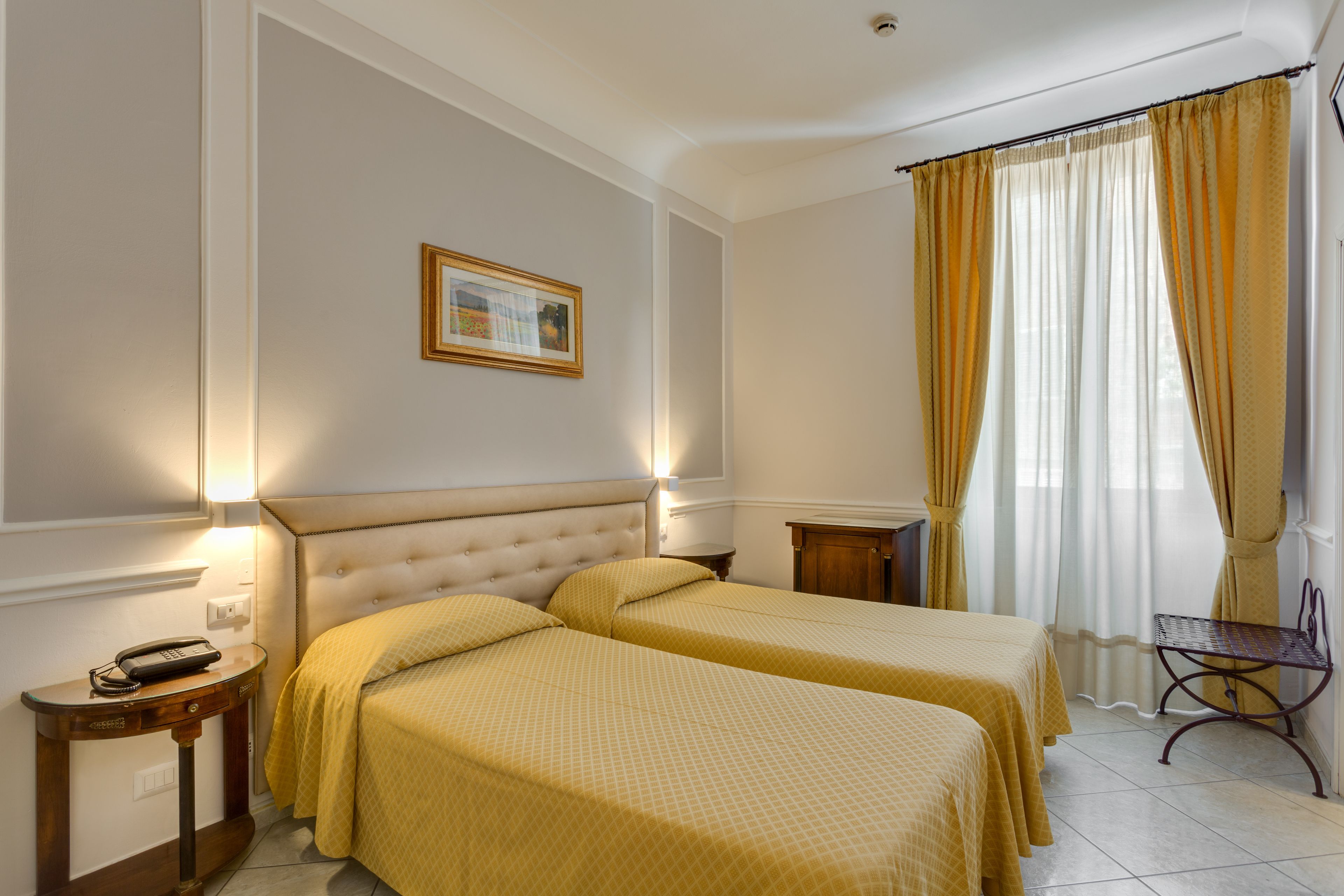 Una habitación de hotel con dos camas, una mesita de noche con teléfono, una pequeña mesa con una silla y una ventana con cortinas.