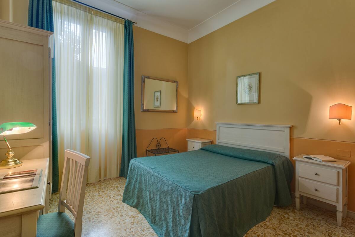 Habitación de hotel con cama, escritorio, silla, mesita de noche, lámpara y ventana con cortinas.