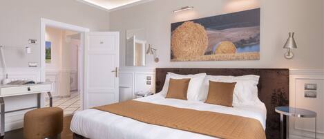 A modern hotel room with a large bed, a desk, and a chair. There is a painting on the wall and a lamp on the side table.