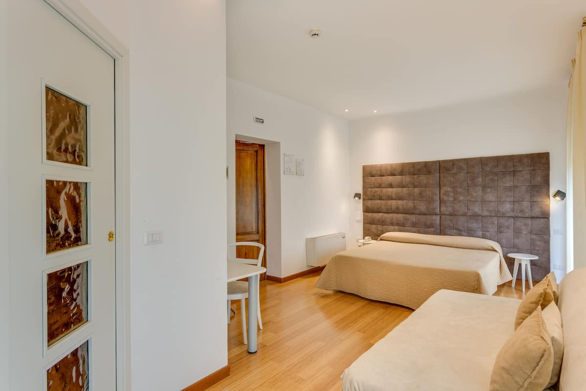 Habitación de hotel con una cama grande, una cama más pequeña, un escritorio y una silla. Se observa una puerta decorada con cuadros y una pared blanca.