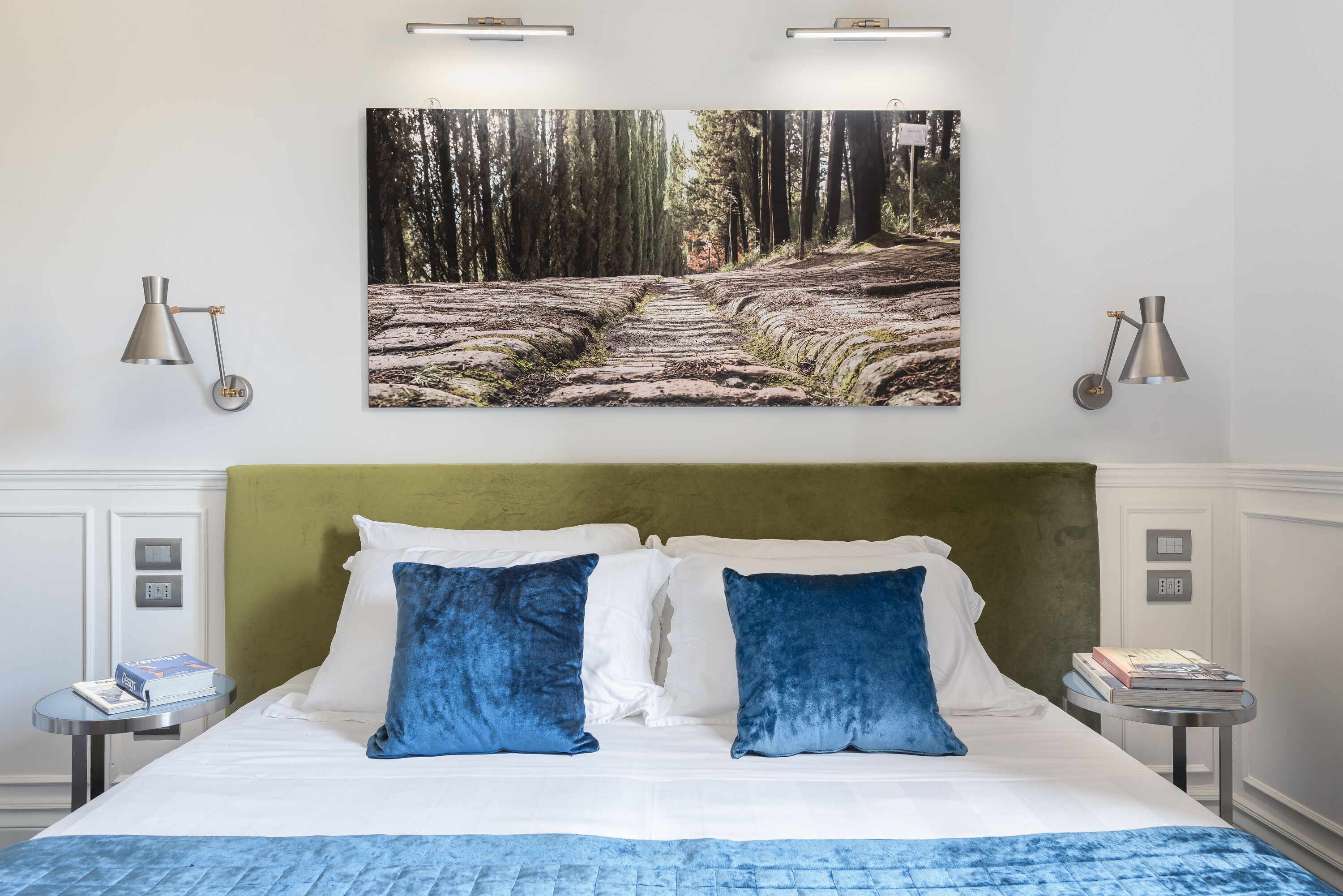Habitación de hotel con una cama, dos almohadas, cabecero, mesita de noche y un cuadro colgado en la pared con un paisaje boscoso.