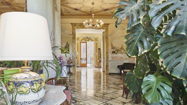 Un vestíbulo con piso de mármol, un candelabro, un mostrador de recepción y grandes plantas verdes.