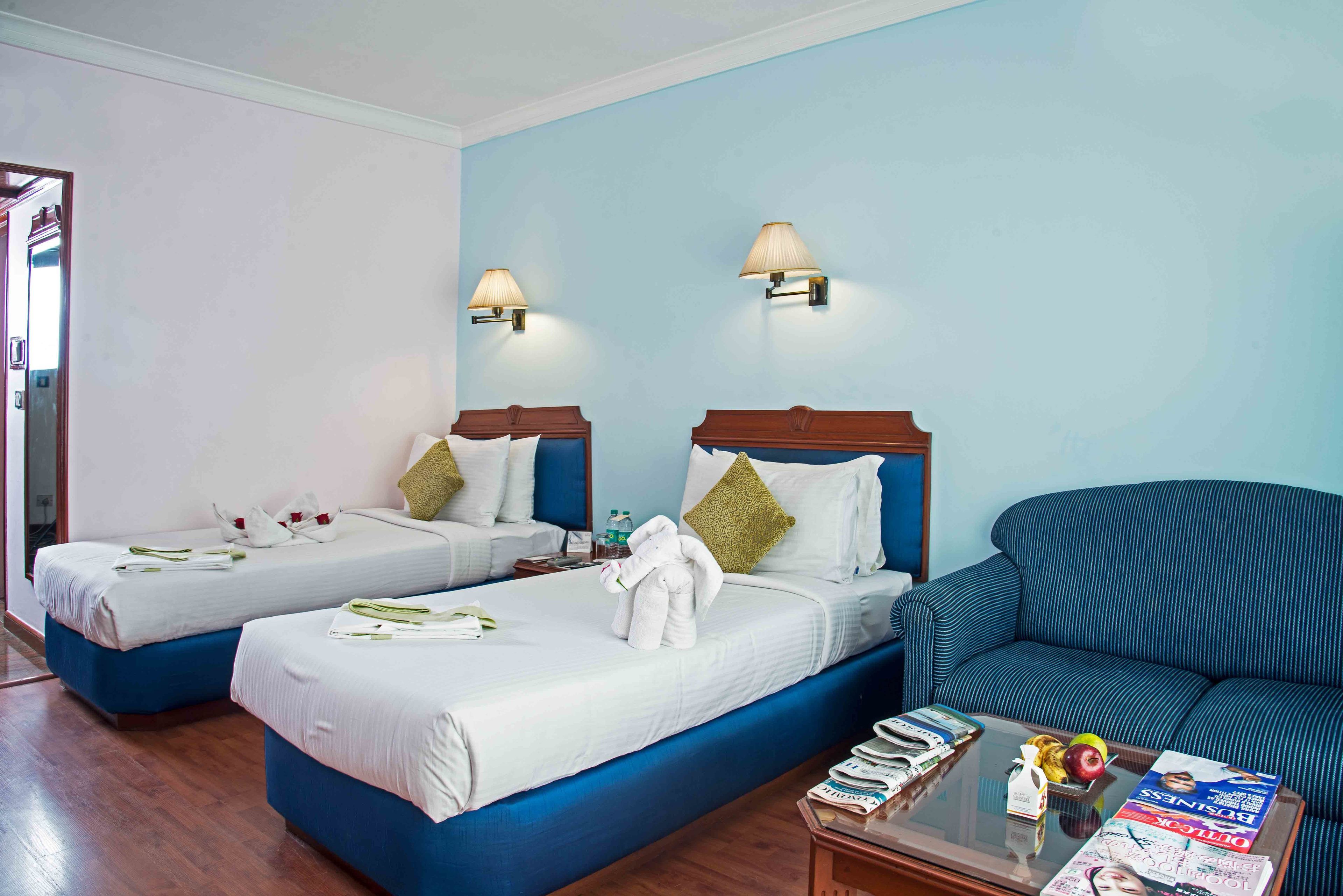 A hotel room with two beds, a blue sofa, and a coffee table with magazines and a fruit bowl.