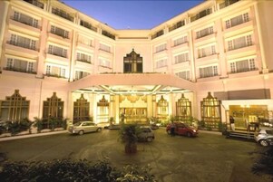 A multi-story hotel with a central courtyard, featuring a grand entrance with a canopy, several balconies, and a well-maintained lawn.