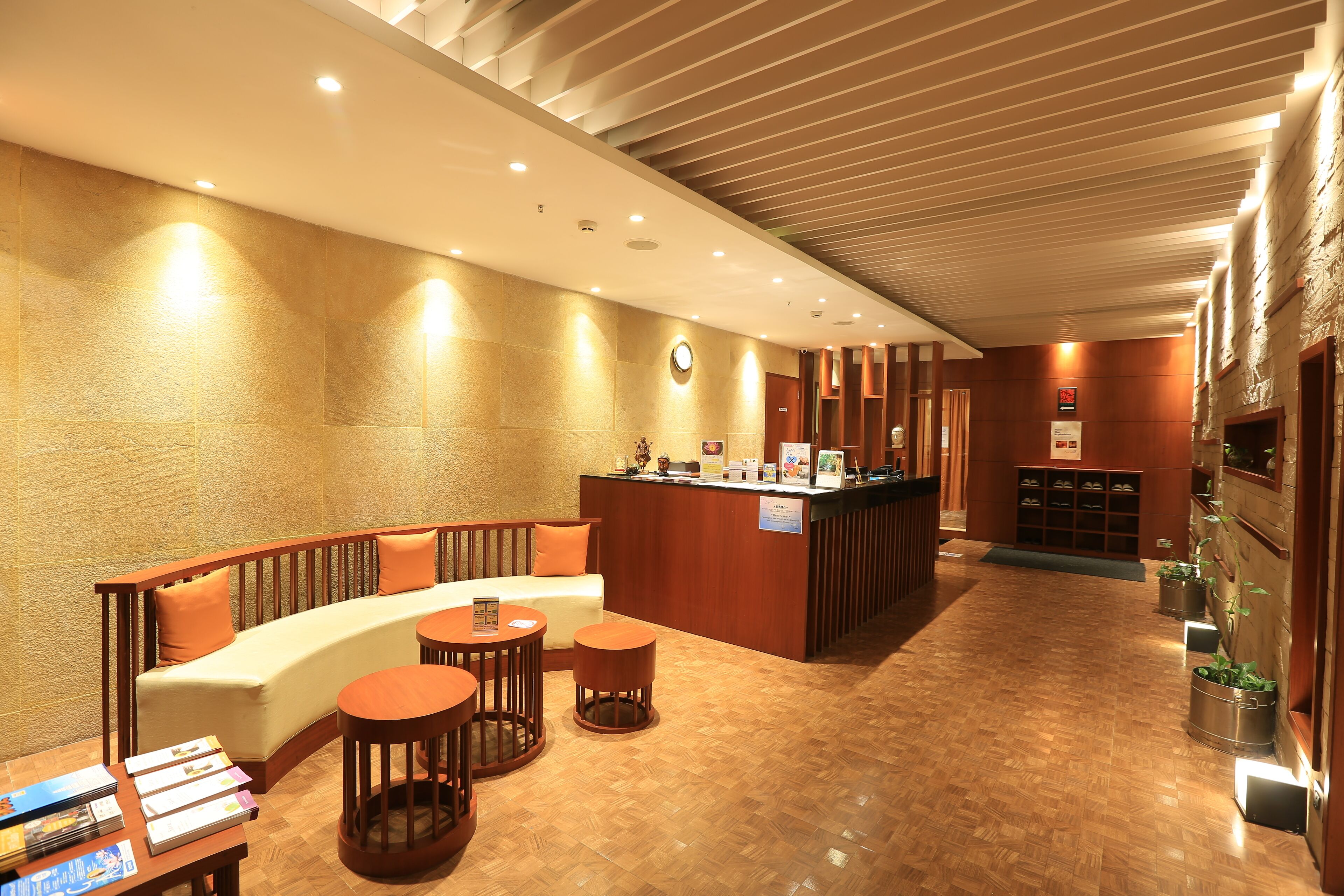 Um hall de hotel com uma receção, uma área de estar com um sofá e duas mesas redondas, e chão de madeira.