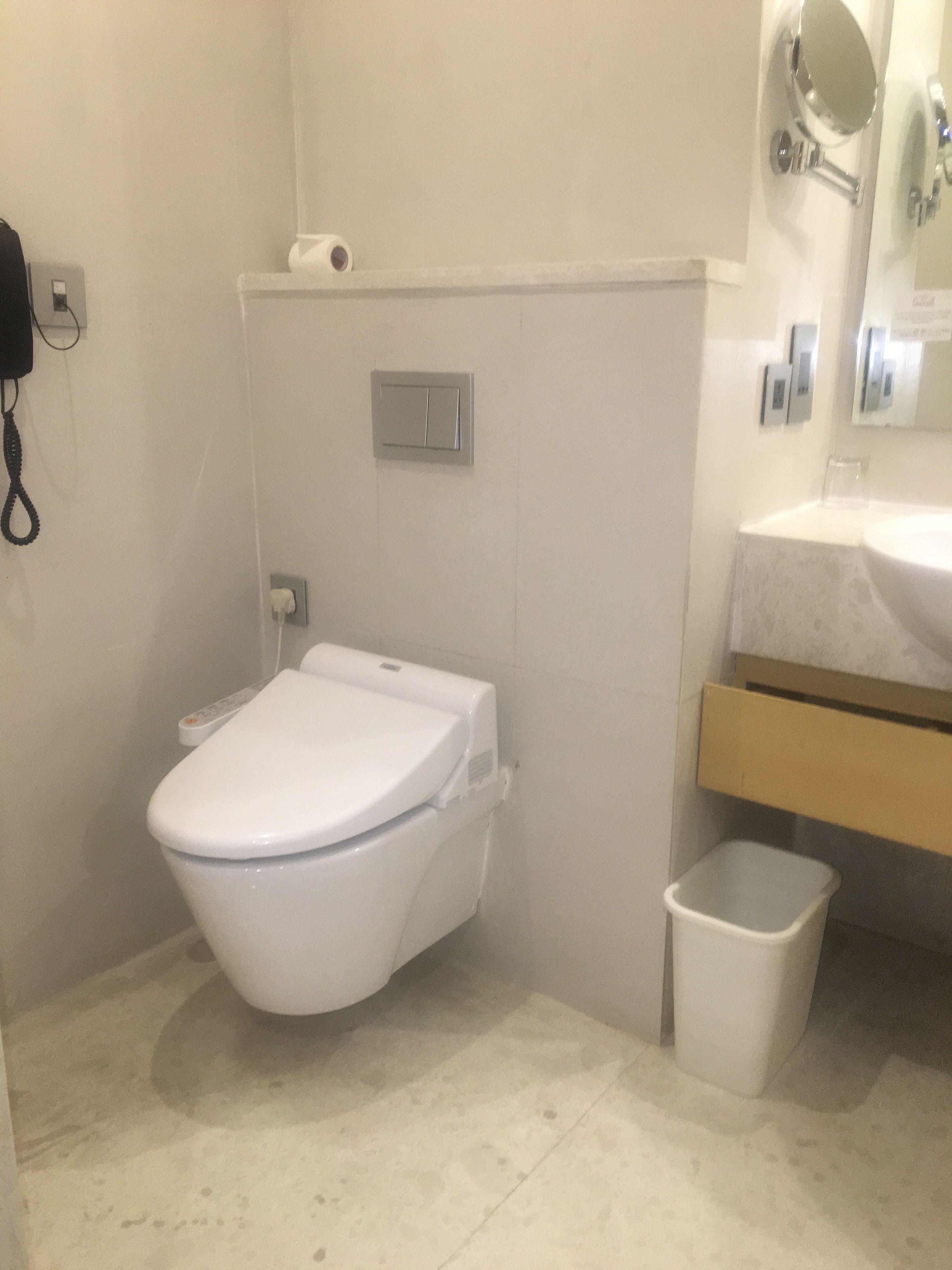 A small bathroom with a toilet, a trash bin, a wall-mounted light switch, and a mirror.