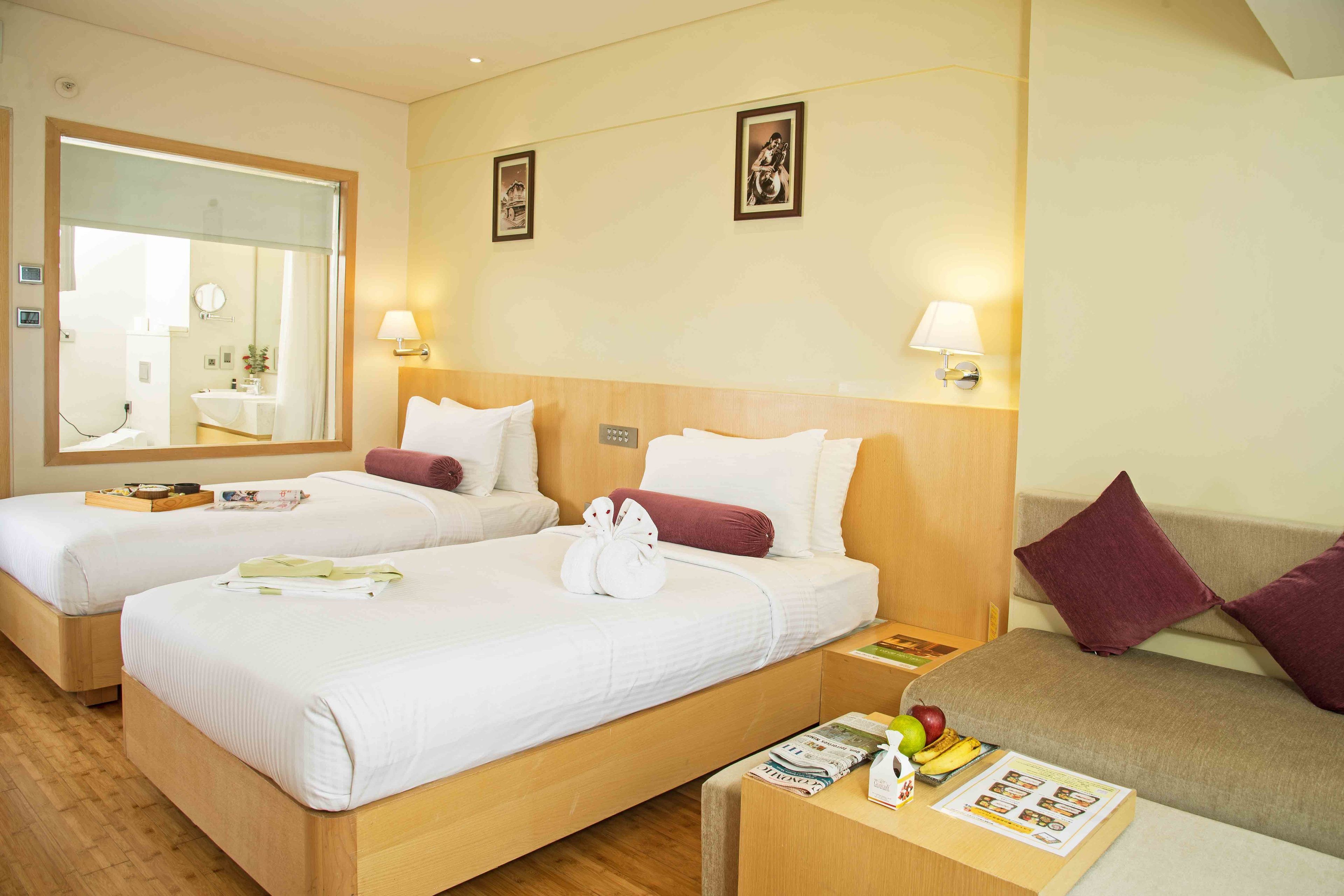A hotel room with two single beds, a wooden headboard, a wooden nightstand with a magazine and fruit, and a sofa with a cushion.