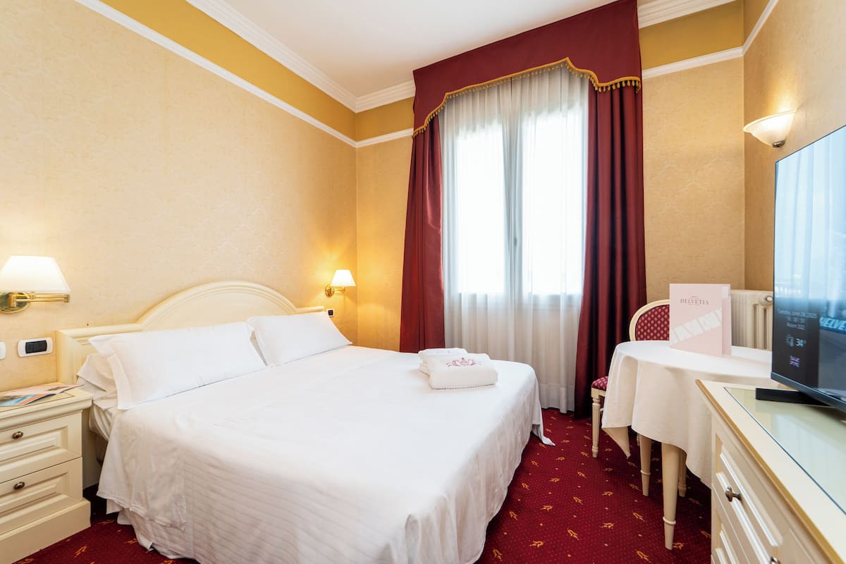 Habitación de hotel con cama, escritorio, televisor, mesita de noche y ventana con cortinas.