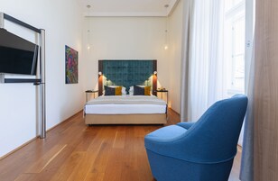 Quarto de hotel moderno com uma cama grande, uma poltrona azul, uma TV de tela plana e um quadro na parede.