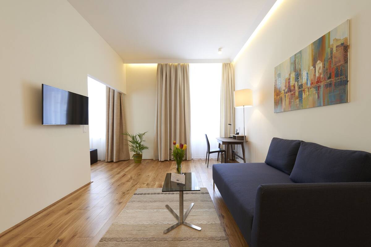 A modern living room with a dark sofa, a flat-screen TV mounted on the wall, a small table with a vase of flowers, and a large abstract painting.