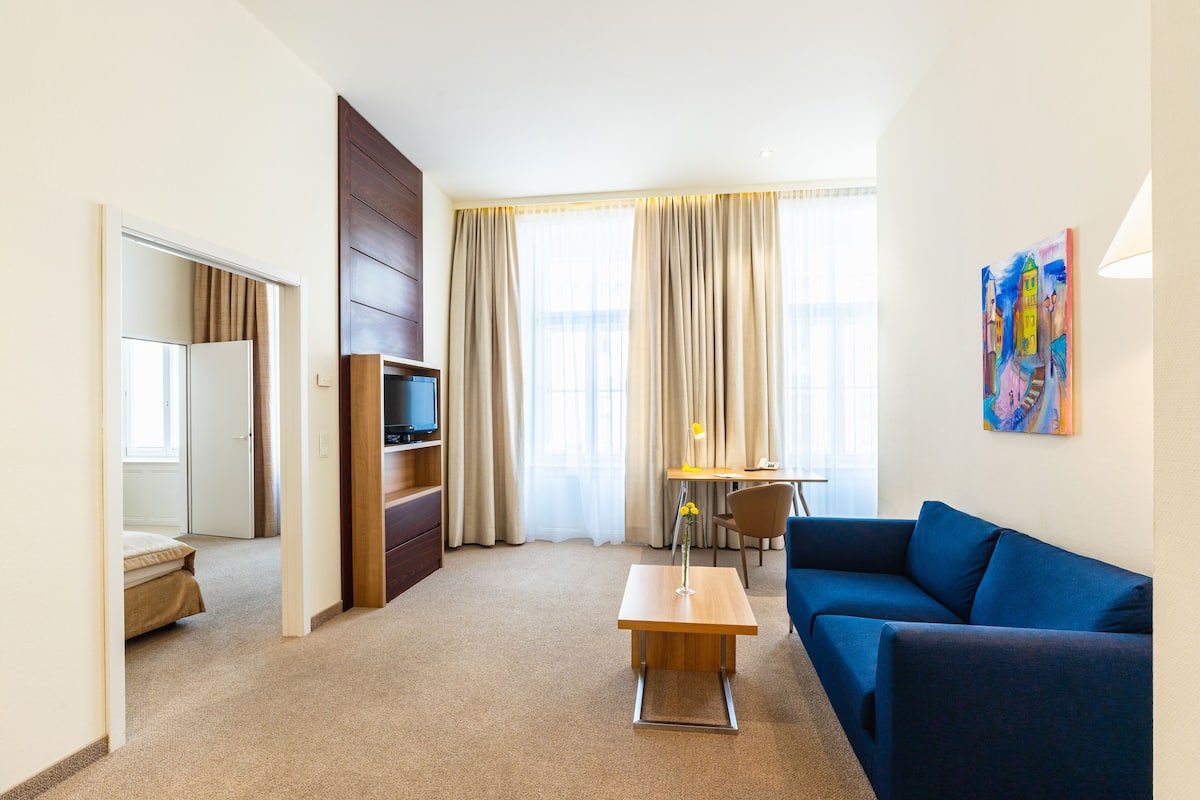 A hotel room with a blue sofa, a wooden coffee table, a TV on a wooden stand, a desk with a chair, and a painting on the wall.