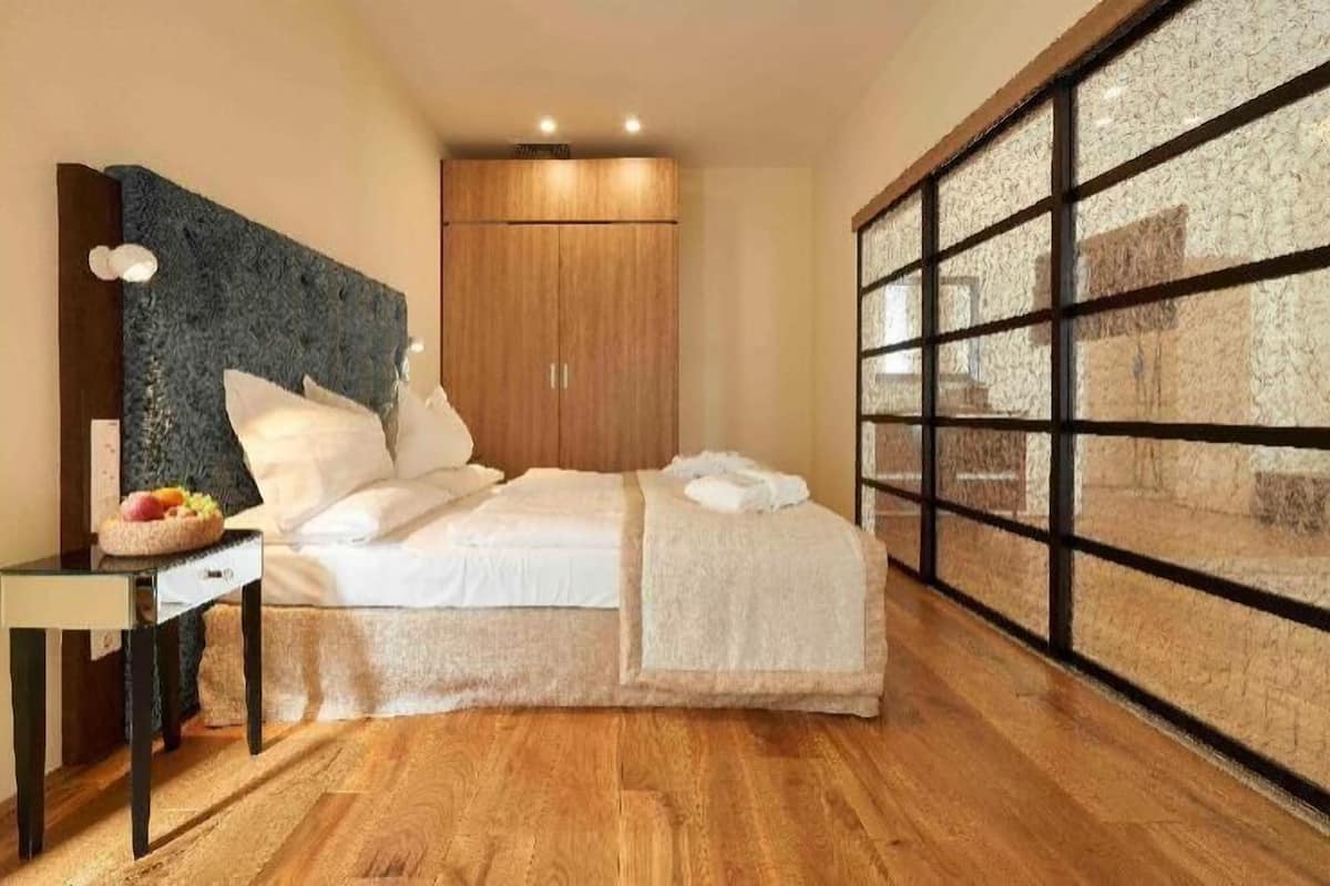 A bedroom with a bed, a desk with a basket of fruit, a wardrobe, and a large window with a view.
