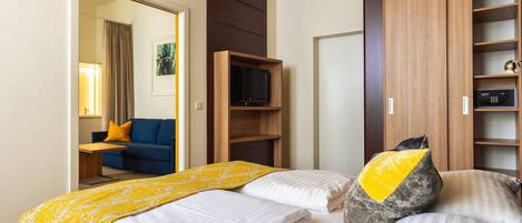 Quarto de hotel com cama, televisão, guarda-roupa e área de estar com sofá e mesa.