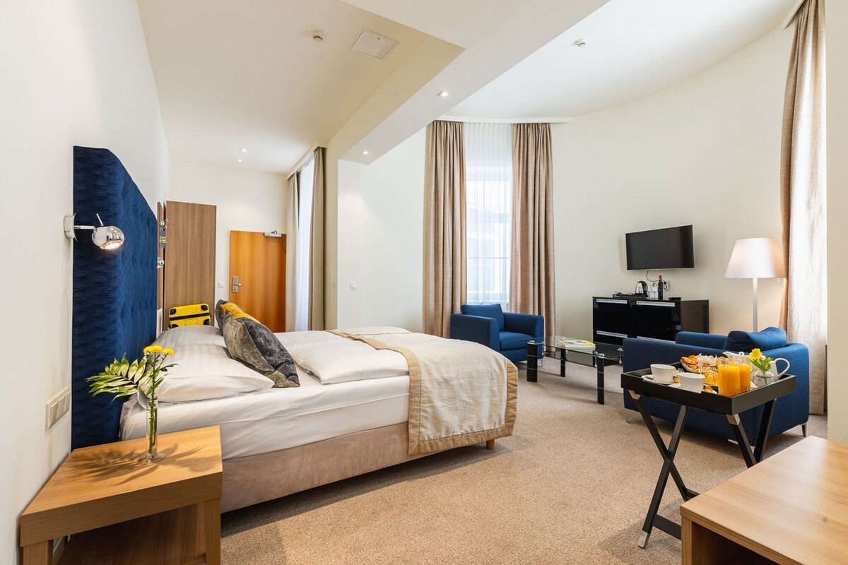 A hotel room with a bed, a desk, a chair, a sofa, a TV, and a breakfast tray.
