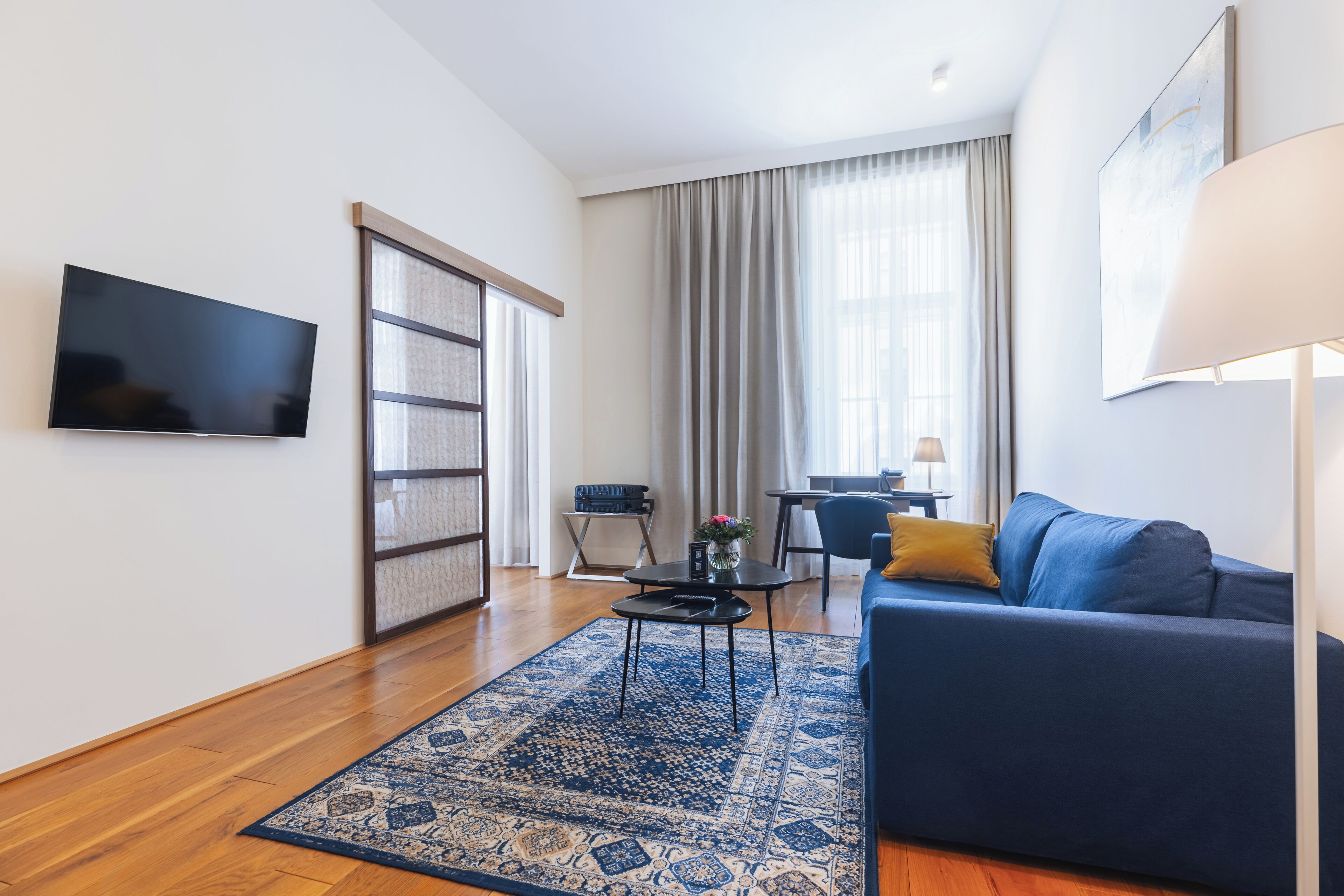 A modern living room with a blue sofa, a flat-screen TV mounted on the wall, a wooden floor, and a large window with curtains.