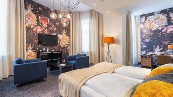 Quarto de hotel com uma cama grande, duas poltronas, uma escrivaninha, televisão de tela plana e papel de parede floral.
