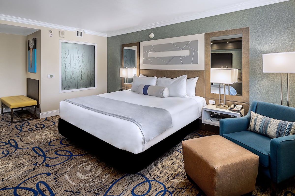 Una habitación de hotel con una cama grande, un sillón azul, una mesita de noche con una lámpara y un banco.