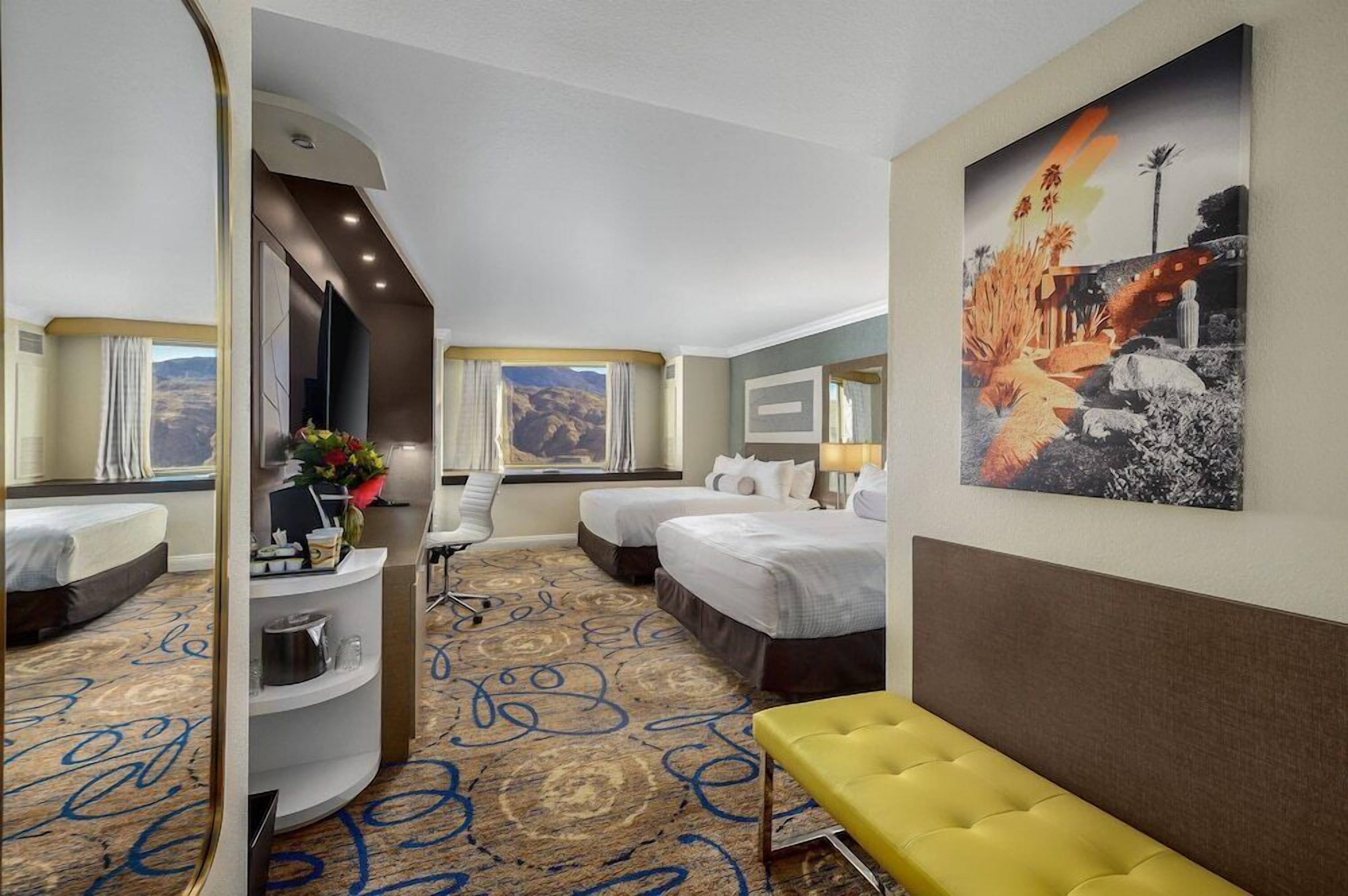 A hotel room with two beds, a desk, a chair, a bench, a large window, and a painting on the wall.