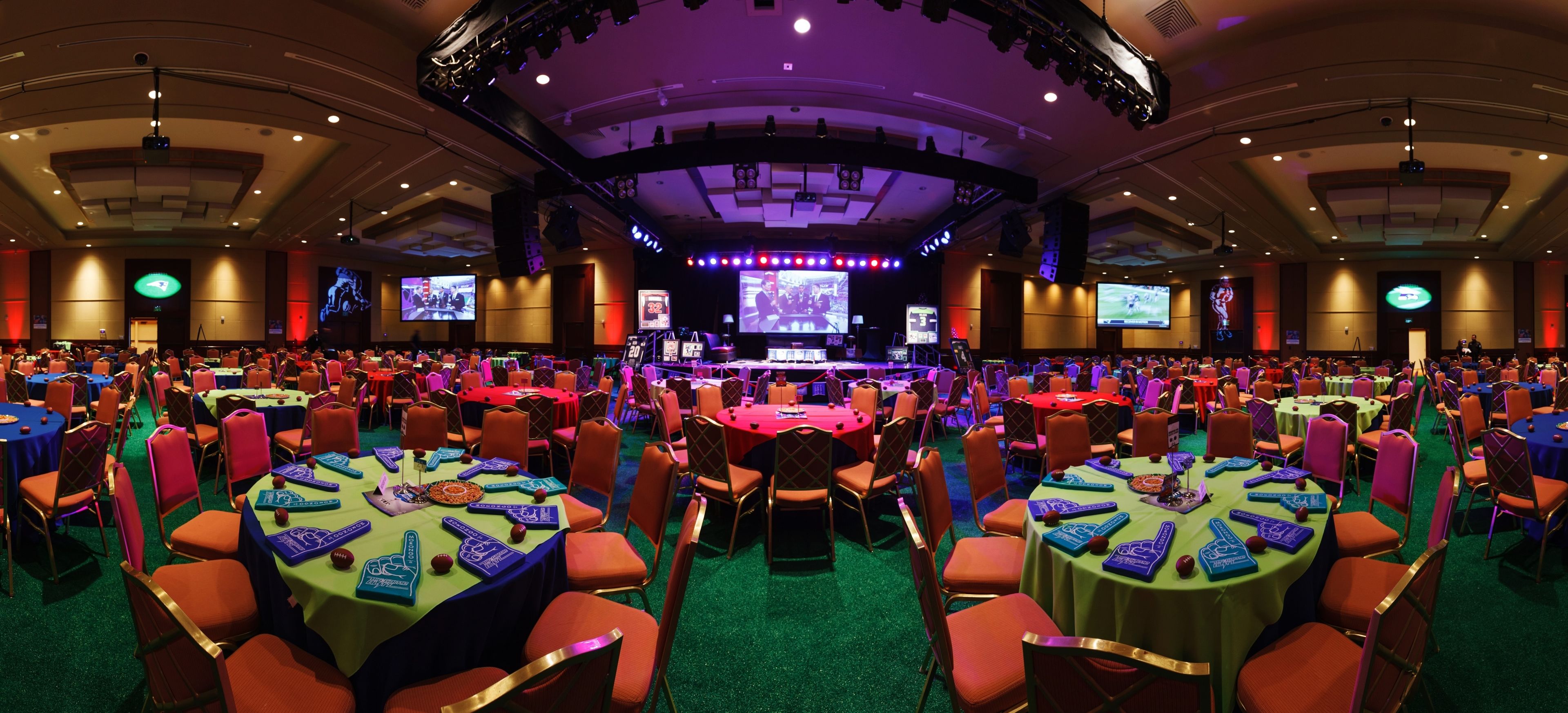 A large conference room with multiple tables and chairs, equipped with screens and a stage for presentations.