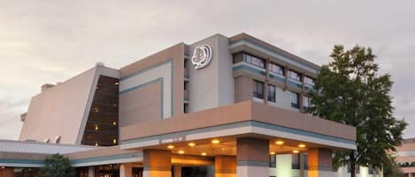 A modern hotel building with a prominent logo, a covered entrance, and a clear sky.