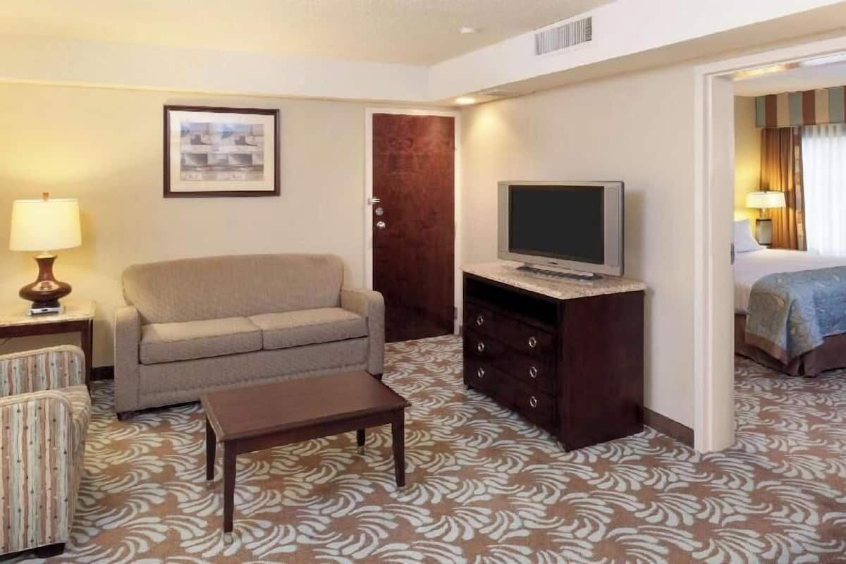 Ein Hotelzimmer mit einer Couch, einem Fernseher auf einem Schrank, einem gemusterten Teppich, einer Tür und einem Bett mit blauer Decke.