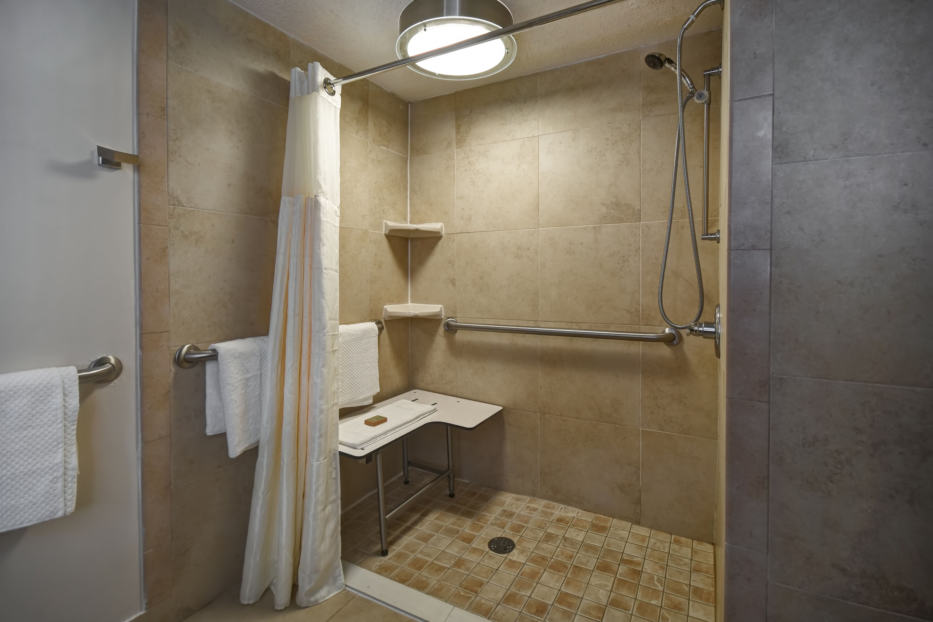 Ducha a ras de suelo con azulejos color crema, un toallero blanco, panel de control de la ducha y cabezal de ducha manual.