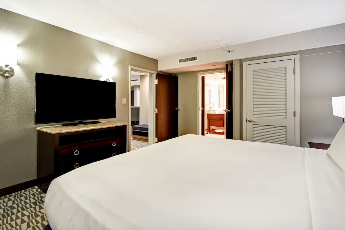 Ein Hotelzimmer mit einem großen Bett, einem Flachbildfernseher, einem Schminktisch und einer Tür zu einem Badezimmer.