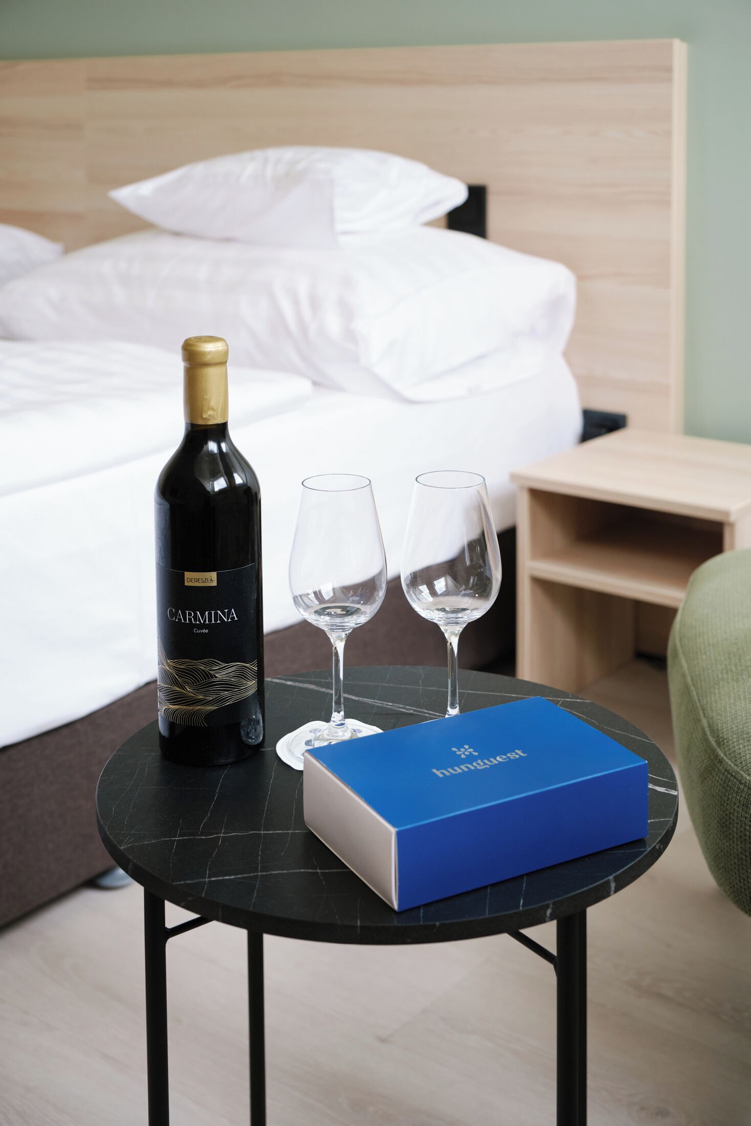 A bottle of Carmina wine, two glasses, and a blue box on a small table in a hotel room.