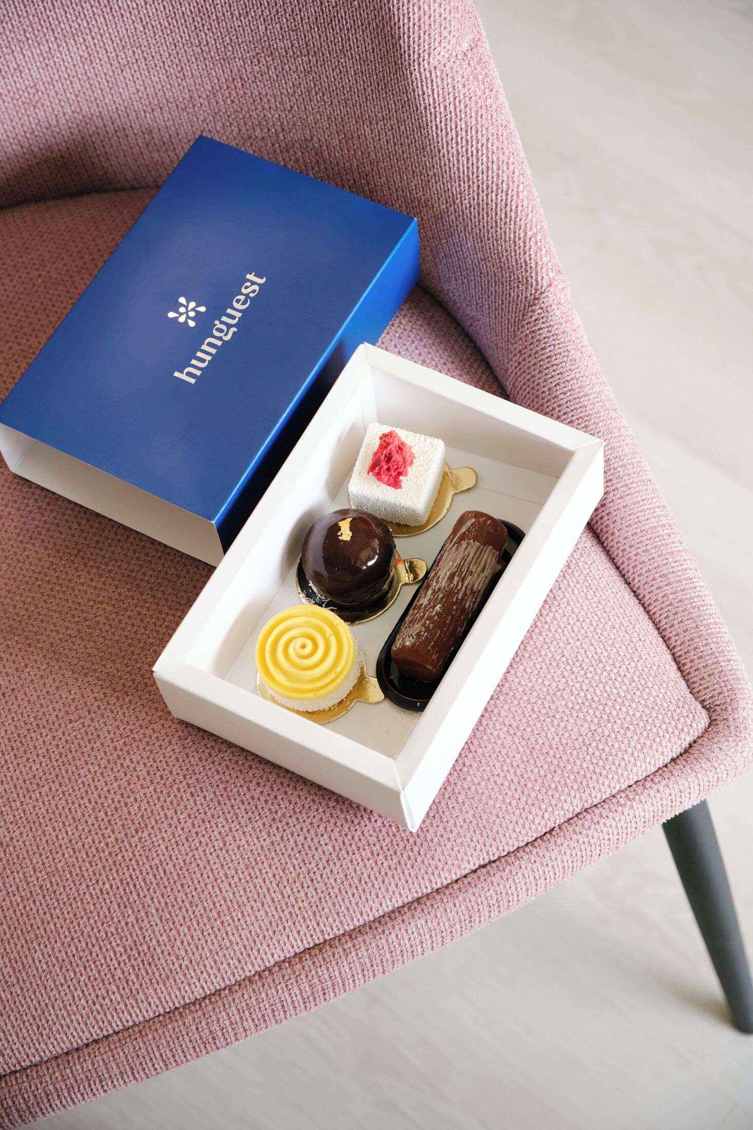 Uma caixa azul com a etiqueta 'Hunguest' sobre uma cadeira rosa, com uma caixa branca contendo diversos doces.
