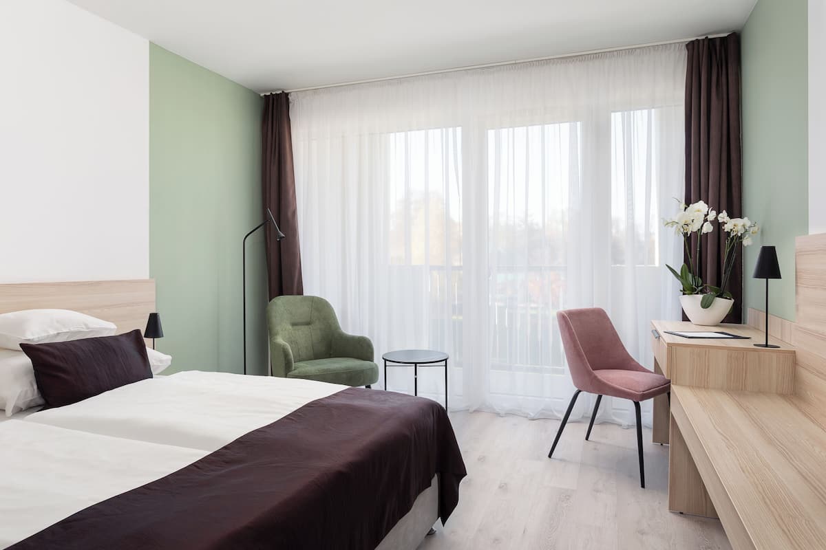 Habitación de hotel con cama, dos sillas, un escritorio, una lámpara y una ventana con cortinas.