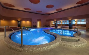 A large indoor swimming pool with a tiled edge, a lounge area with chairs, and a clock on the wall. - Hunguest Hotel Flora (Eger)