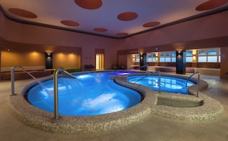 Una amplia piscina interior con borde de azulejos, un área de descanso con sillones y un reloj en la pared.