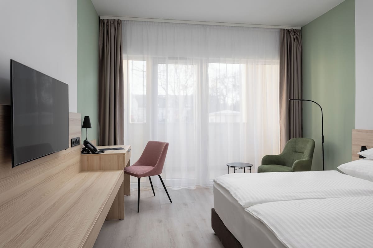 Habitación de hotel con cama, escritorio con computadora, silla, televisor y ventana con cortinas.