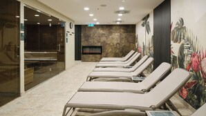 A room with a row of sunbeds, a glass partition, a wall-mounted TV, and a mural with tropical plants. - Hunguest Hotel Flora (Eger)