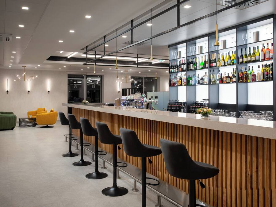 Un bar moderno con mostrador de madera, taburetes negros y una variedad de botellas y vasos en estantes.