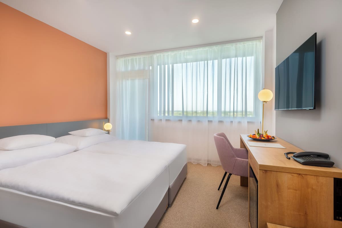 Quarto de hotel com uma cama grande, uma escrivaninha com cadeira, uma televisão de tela plana e uma janela com cortinas translúcidas.