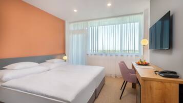 Habitación de hotel con una cama grande, un escritorio con silla, un televisor de pantalla plana y una ventana con cortinas ligeras.