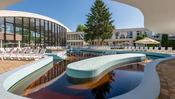 Una piscina con sillas de descanso, un edificio moderno con grandes ventanales y un árbol al fondo.