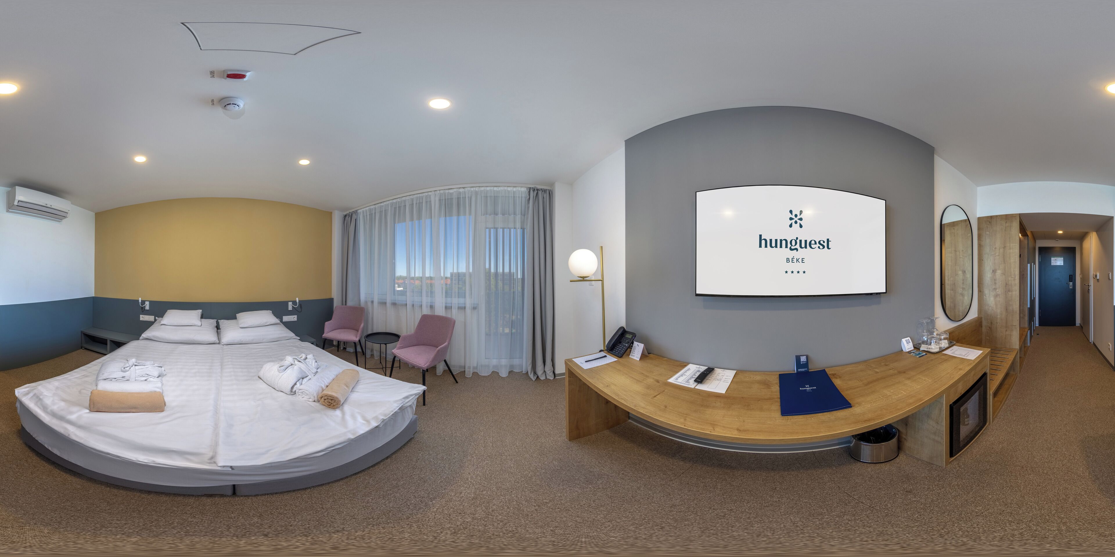 A hotel room with a bed, a desk, a chair, a reception desk, and a large window.