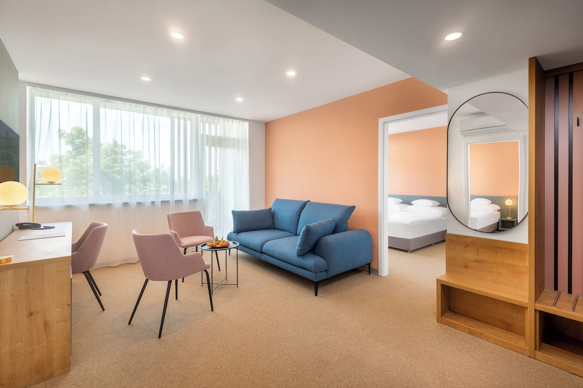 Quarto de hotel moderno com um sofá azul, uma escrivaninha de madeira, uma mesinha com frutas e uma janela grande com cortinas leves.