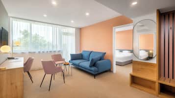 A modern hotel room with a blue sofa, a wooden desk, a small table with fruit, and a large window with sheer curtains.